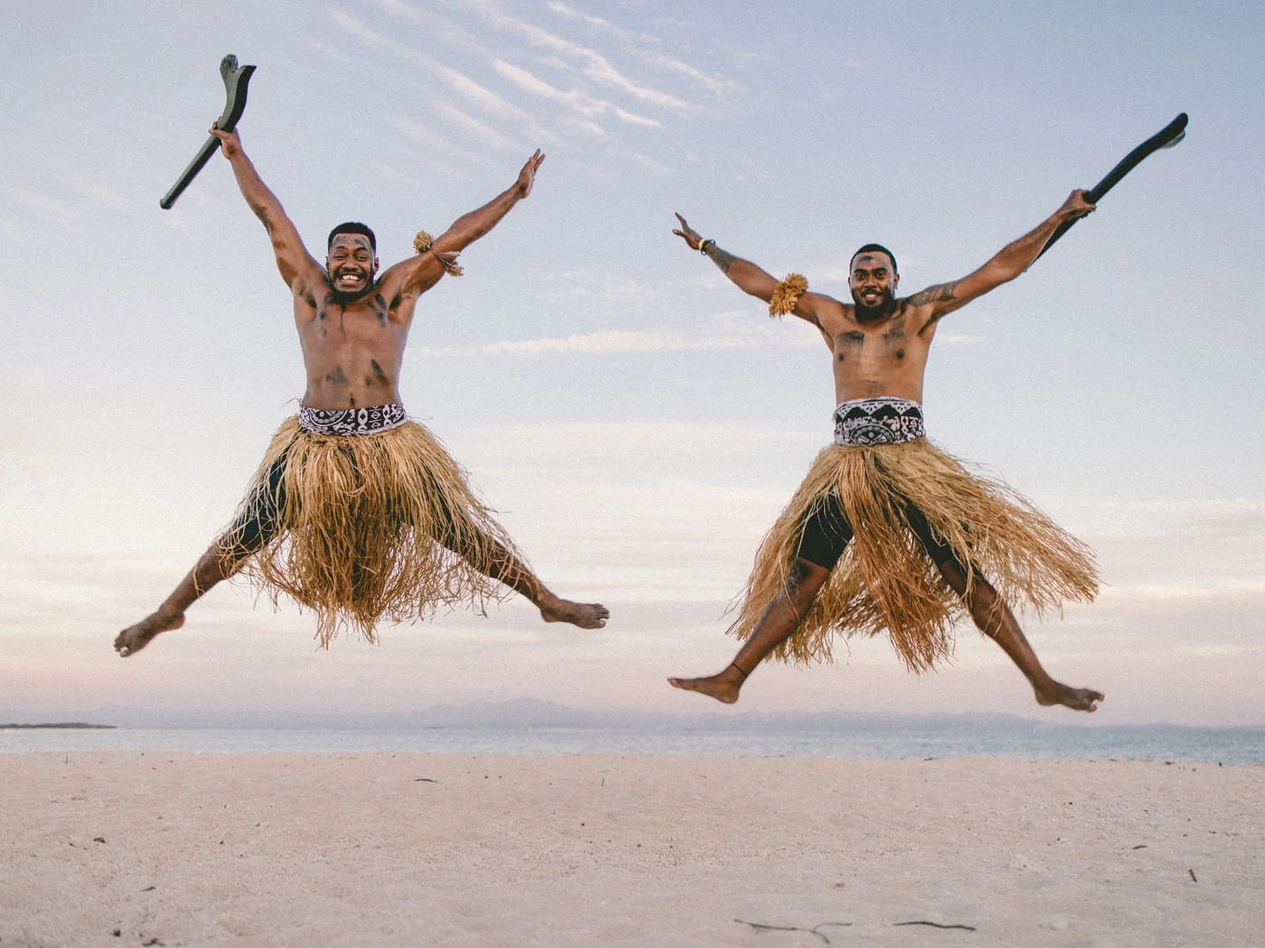 Two men in traditional Fijian grass skirts leaping on a beach, clubs raised overhead